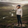 Femme debout sur un rocher dans une vallée écossaise, portant une casquette Baker Boy Harris Tweed® bleu marine avec un pull crème.
