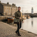 Un homme en veste de tweed portant la grande sacoche Harris Tweed® à motif chevron marron-vert à l'épaule, marchant le long d'un port de pêche pittoresque avec un phare en arrière-plan.