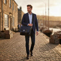 Homme élégant en costume bleu portant la grande sacoche Harris Tweed® au motif chevron bleu marine en bandoulière, marchant sur des pavés dans un décor portuaire au soleil couchant.