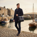 Homme en caban bleu marine portant la grande sacoche Harris Tweed® au motif tartan bleu et gris en bandoulière sur un quai de port, illustrant un look marin classique et urbain.