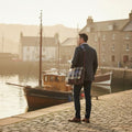 Homme de dos portant la grande sacoche Harris Tweed® au motif tartan bleu, gris et marron en bandoulière, marchant dans un port historique face à des bateaux de pêche traditionnels.