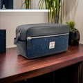 Grande trousse de toilette Harris Tweed® au motif chevron bleu marine posée sur un plateau en bois sombre, mettant en avant le contraste entre le coloris du tweed et le cuir végan noir.
