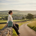 Une femme assise de profil sur un muret en pierre dans un paysage de campagne verdoyant, portant le petit sac à dos Harris Tweed® au motif chevron turquoise bleu-vert.
