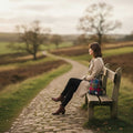 Une femme assise sur un banc en bois le long d'un chemin de campagne, le petit sac à dos Harris Tweed® au tartan multicolore bleu, rose, rouge et vert posé à ses côtés.