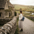 Une femme debout de dos devant un muret en pierre dans un village historique, portant le petit sac à dos Harris Tweed® aux tons violet et bleu turquoise.