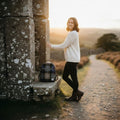 Une femme debout à côté d'une structure en pierre au soleil couchant, portant le petit sac à dos Harris Tweed® au tartan marron, bleu et gris.