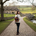 Une femme marchant sur un chemin de campagne devant un cottage, portant le petit sac à dos Harris Tweed® au tartan traditionnel rouge, bleu et jaune.