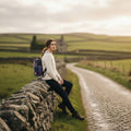 Une femme assise sur un muret en pierre dans un paysage vallonné, portant le petit sac à dos Harris Tweed® tartan aux nuances de rose fuchsia et bleu.