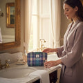 Une femme en peignoir mauve préparant ses produits de soin à côté de sa petite trousse de toilette Harris Tweed® tartan bleu, vert et violet dans une salle de bain lumineuse.