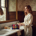 Une femme rousse en peignoir de satin devant un miroir de salle de bain utilisant la petite trousse de toilette Harris Tweed® au tartan traditionnel bleu, rouge et jaune.