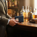 Homme en veste de tweed tenant le portefeuille deux volets Harris Tweed® motif à chevron bleu marine et noir devant un comptoir, avec des bouteilles de spiritueux en arrière-plan.