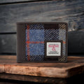 Portefeuille 3 volets Harris Tweed® posé sur une planche de bois rustique, mettant en valeur la texture de la laine au motif tartan bleu, gris et marron et les finitions en cuir végan marron.