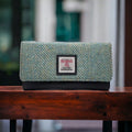 Portefeuille enveloppe Harris Tweed® posé sur un bureau en bois vintage, mettant en avant le tissage chevron bleu-vert et l'étiquette Orb Mark.