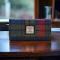 Portefeuille enveloppe Harris Tweed® au tartan bleu, rouge, vert et violet posé sur une structure en bois rustique, soulignant le contraste du tartan coloré.