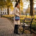 Sac bandoulière carré Harris Tweed® aux tons bleu, vert et violet porté sur un trench beige dans un parc en automne.