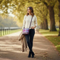 Sac bandoulière carré Harris Tweed® aux tons rose, bleu et violet, porté sur un pull crème et un pantalon noir dans un parc en automne.
