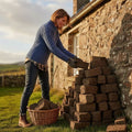Femme empilant des blocs de tourbe devant une maison en pierre, vêtue d'un pull jumper en tricot de laine mérinos bleu inspiré des traditions d'Irlande et d'Aran.