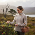 Femme vêtue du pull Haymoor gris chiné en tricot de laine mérinos de style Aran, cueillant des baies sauvages dans la lande d'Irlande, un panier en osier à la main.