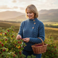 Femme vêtue du pull jumper Haymoor en tricot de laine mérinos, cueillant des baies au coucher du soleil dans la campagne d'Irlande, avec des montagnes en arrière-plan, style Aran.