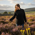 Femme cueillant des branches de bruyère dans la lande d'Irlande, vêtue d'un pull jumper noir Merston au tricot Aran authentique en laine mérinos et col montant.