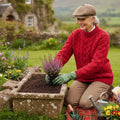 Femme mûre jardinant dans un potager en Irlande, portant un pull jumper Merston rouge vif au tricot artisanal en laine mérinos fidèle au style Aran et col montant.
