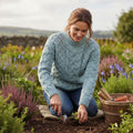 Femme souriante jardinant au milieu des fleurs, vêtue du pull jumper bleu clair Merston, un tricot de laine mérinos robuste et élégant typique du savoir-faire d'Irlande et d'Aran.
