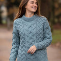 Portrait d'une femme joyeuse portant le pull jumper Merston en coloris bleu clair, soulignant la texture riche d'un tricot Aran authentique en laine mérinos originaire d'Irlande.