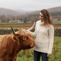Femme vêtue du pull jumper Whitmore écru au tricot en laine mérinos robuste de style Aran d'Irlande, caressant une vache Highland dans un paysage bucolique d'Écosse.