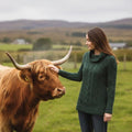 Femme portant le pull jumper Whitmore en tricot de laine mérinos vert forêt, un modèle Aran traditionnel d'Irlande, caressant une vache Highland dans un pâturage.