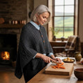Femme vêtue du poncho Remington en tricot de laine mérinos gris anthracite préparant des légumes dans une cuisine rustique d'Irlande, illustrant le style Aran chaleureux.